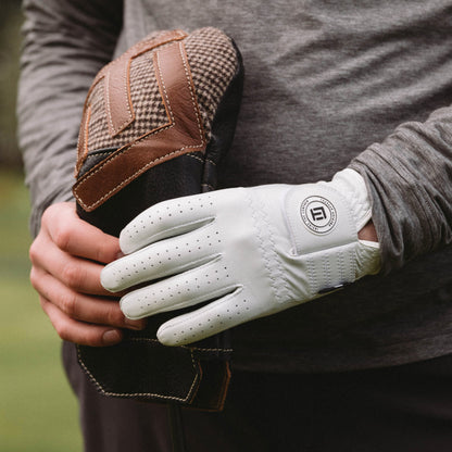 Close-up of a person wearing a white golf glove with a brand logo, holding a brown and black glove.